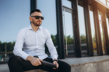 Young successful businessman working on laptop, bearded man in business suit looking at camera and smiling, talking on the phone near a modern office center outsideの写真素材
