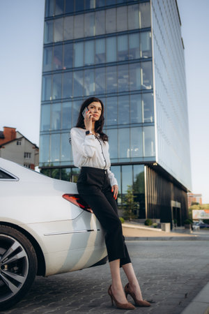 Business Woman Uses Smartphone While Leaning on Her Premium Class Car. Big City with Skyscrapers in the Backgroundの写真素材