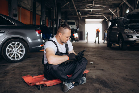 Young professional technician car mechanic man 20s in blue overalls white t-shirt use hold mobile cell phone browsing fix problem with raised hood bonnet work in vehicle repair shop workshop indoor.の写真素材