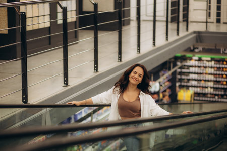 Portrait of smiling pretty young woman holding on escalator handrail and riding escalator going up in shopping mall, looking away, paper bags with purchases in hands, blurred background. high quality photoの写真素材