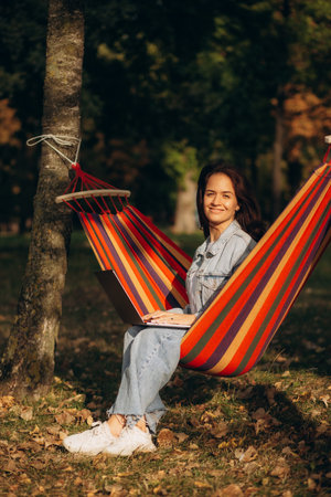 Beautiful young woman browsing Internet on laptop while lying in hammock, picturesque tropical park...