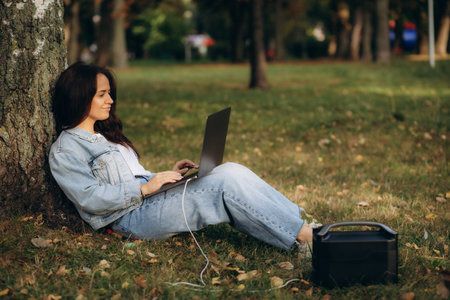A woman works on a laptop under a tree and charges it from a portable charging station
