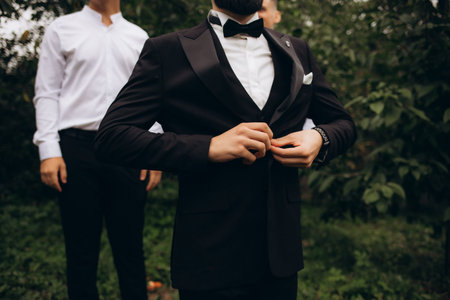 The groom in a suit and tie is standing on the background of greenery, on the bachelor's jacket is a...
