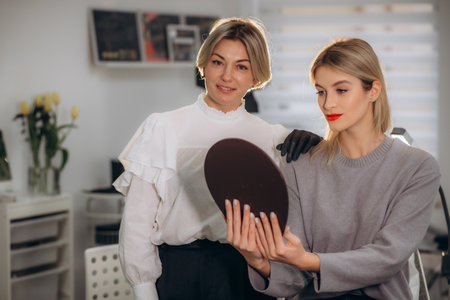 Bottom view of mirror reflection of smiling caucasian girl looking at her beautiful lips while female beautician looking at her after permanent makeup lips procedure in beauty salon. Beauty concept.の写真素材