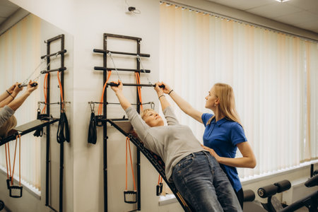 Positive senior woman doing stretching exercises on Pilates reformer as part of injury rehabilitation course. Therapeutic physical training conceptの写真素材