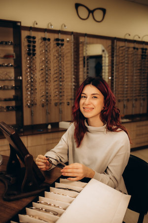 Woman chooses glasses in the store. Blonde in a beige sweater measures glasses near the mirror. Girl on a background of shop windows with different models of glasses. High quality photoの写真素材