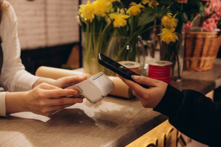 Cropped shot of female florist holding smartphone paying for flowers in flower shop