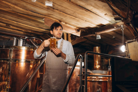 Expert brewer in apron holds glass of craft beer and checking quality and color. Worker man...