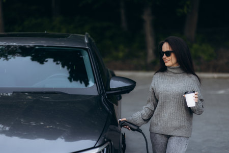 Happy young adult woman smiling wide, looking away, charging automobile battery from small public station, standing near electric car, drinking coffee and talking on smartphoneの写真素材
