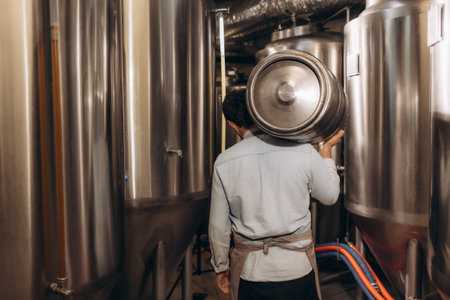 Shot of young man with metal beer barrels at brewery. Brewer carrying keg at brewery plant.