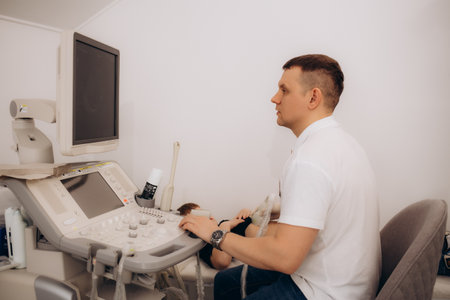 Charming young male doctor smiling to the camera, sitting at his office near ultrasound scanning machine. Handsome friendly doctor enjoying working at the hospital, copy space. Ultrasonic conceptの写真素材