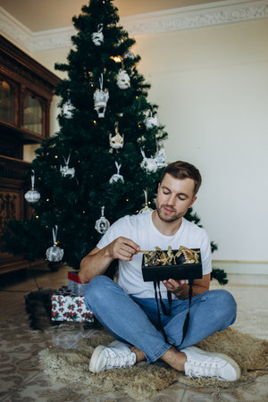 Portrait of happy handsome young man holding cardboard box with green ribbon near Christmas tree and looking at camera.の写真素材