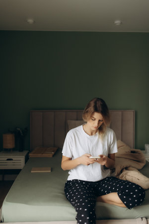 A blonde woman sits on the bed texting on her phoneの写真素材