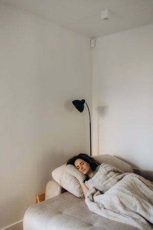 Full length peaceful woman resting under warm blanket on cozy couch in living room, calm young female with closed eyes sleeping, taking nap or daydreaming, lying on soft pillow with hands under cheekの写真素材