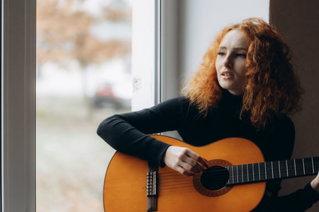 A redheaded girl plays guitar by the windowの写真素材