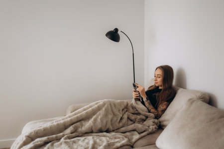 Image of happy nice woman in pajamas smiling and using cellphone while sitting on sofa at living roomの写真素材