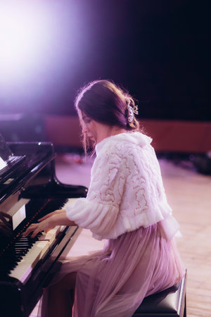A young woman elegantly dressed in a soft gown skillfully playing a black grand piano under ambient lighting, radiating a serene and artistic atmosphere in a sophisticated setting.の写真素材
