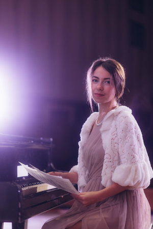 A poised woman with refined attire sits at a grand piano holding sheet music, with soft, warm lighting highlighting her serene expression and graceful demeanor, evoking a sense of musical artistry and elegance.の写真素材