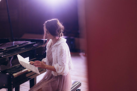 A young woman in a light outfit reviews sheet music while sitting at a grand piano.の写真素材