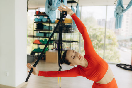 A woman wearing a red outfit is stretching with fitness equipment in a bright studio. The image...