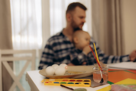 A father holding his baby while painting and coloring eggs at a table filled with art supplies. The warm light and nurturing interaction evoke a sense of familial creativity and care.の写真素材