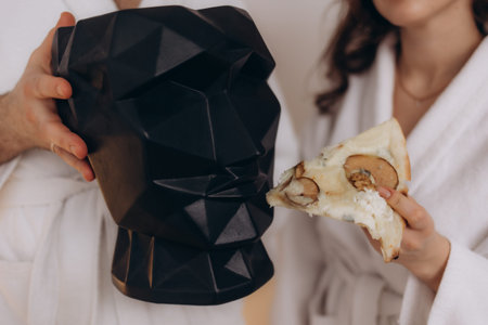 An artistic black geometric head sculpture and a slice of pizza held by people in white robes,...