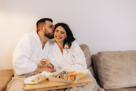 A joyful couple in white robes share sushi, expressing love and relaxation in a cozy environment. They exude happiness and connection, seated on a living room sofa with a sushi platter.の写真素材