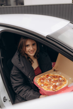 A smiling young woman sits in her car holding a delicious pizza in a delivery box, ready to enjoy...
