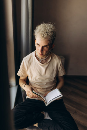 A young man sits by a window indoors, holding and reading a book, surrounded by calm light.の写真素材