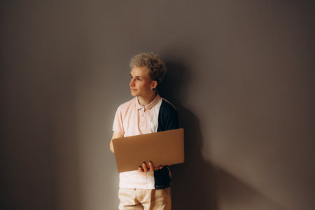 A person is standing against a plain wall, holding a laptop and gazing thoughtfully into the distance, embodying a moment of inspiration or contemplation.の写真素材