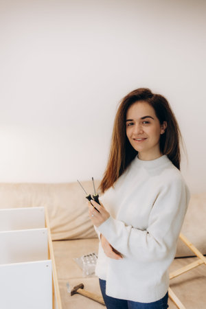 A young woman in a white sweater holds screwdrivers and smiles while assembling furniture indoors....