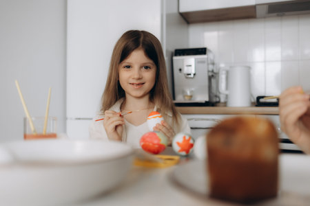 Smiling girl decorating colorful Easter eggs with paintbrushes in a cozy kitchen setting, showcasing...
