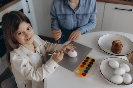 A happy young girl and an adult engaging in egg painting together at a kitchen table, featuring colorful paint trays and traditional holiday decorations creating a joyous and festive atmosphere.の写真素材