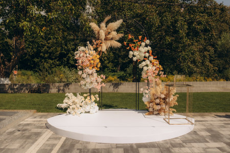 Outdoor wedding ceremony arch adorned with vibrant flowers, pampas grass, and elegant decor, set against a lush green backdrop, creating a perfect romantic setting for a special occasion.の写真素材