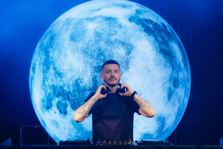 A DJ standing before a magnificent full moon projection, sets an engaging ambiance at a music event,...