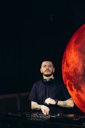 A DJ stands behind a music console, performing under vivid lighting with a dramatic red planetary backdrop, creating a striking and captivating scene of live music entertainment.の写真素材