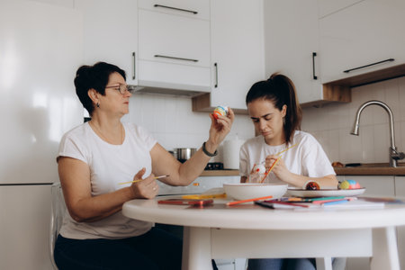 A mother and daughter enjoy painting Easter eggs together at the kitchen table, engaging in a beautiful display of creativity and bonding, surrounded by colorful paints and a bright home environment.の写真素材