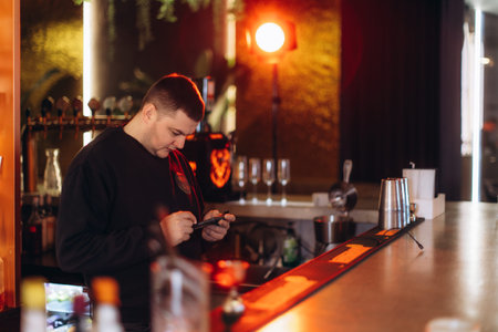 A bartender is focused on using their smartphone while standing behind a bar in a stylish...