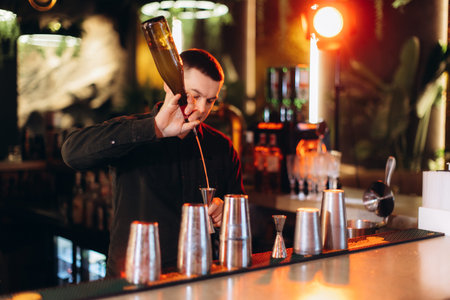 Professional bartender skillfully measures and pours ingredients for a cocktail under soft orange...