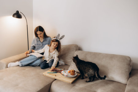 A mother and young daughter engage in creative activities together on a cozy sofa while a curious...