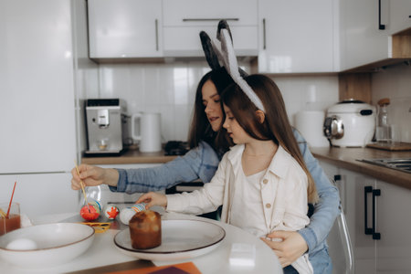A mother and her young daughter, both wearing bunny ears, enjoy painting colorful Easter eggs together in a bright and modern kitchen, sharing a joyful and festive holiday moment.の写真素材