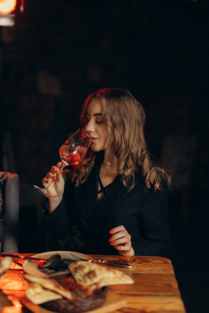 A woman relaxes at a wooden table, enjoying a glass of wine amidst a warm atmosphere, surrounded by delicious food and soft lighting, evoking feelings of charm, taste, and luxury.の写真素材