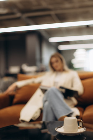 A cozy coffee shop scene featuring a cup of espresso in the foreground with a blurred background including a woman reading comfortably on a fashionable sofa.の写真素材