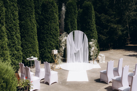 Stunning outdoor wedding venue decorated with a white arch, draped fabric, and elegant chairs, creating a serene and sophisticated ambiance for a special celebration.の写真素材