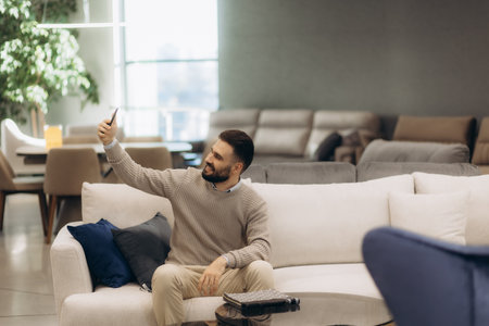 A man sitting on a stylish sofa taking a selfie in a bright and modern living room environment.の写真素材