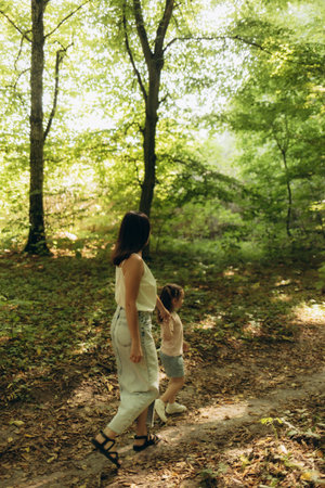 A heartwarming scene of a mother and daughter holding hands while strolling through a lush green forest trail, enjoying quality time and bright sunlight filtering through the trees.の写真素材