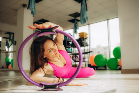A woman in a vibrant sports outfit uses a pilates ring for fitness exercises in a bright studio with gym equipment in the background.の写真素材