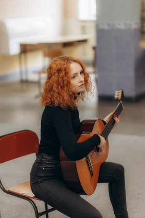 A young woman with curly red hair sits indoors playing an acoustic guitar in a relaxed setting. The...