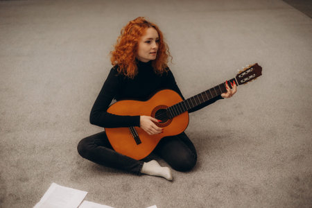A red-haired woman sits on a carpet, strumming an acoustic guitar, surrounded by papers, evoking a...