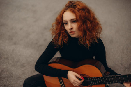 A young woman with vibrant red hair plays an acoustic guitar while sitting indoors, exuding a sense...
